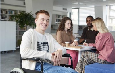Portrait of smiling red haired boy using wheelchair studying with group of students in college library and looking at camera