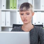 Girl with short blond hair is sitting at her workplace and looking at laptop screen.