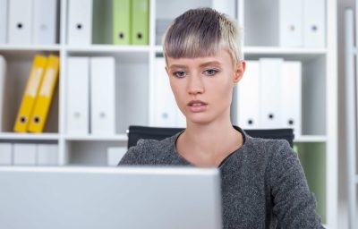 Girl with short blond hair is sitting at her workplace and looking at laptop screen.