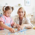 Young mother helping to her daughter with down syndrome collecting puzzles at the table in the room