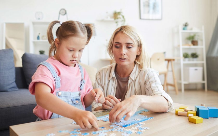Young mother helping to her daughter with down syndrome collecting puzzles at the table in the room