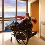 Father kneeling next to disabled son in wheelchair looking out hospital window at ocean and cliffs outdoors.