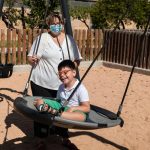 Child with disability in swing with mother pushing, wears blue mask of pandemic coronavirus protection.