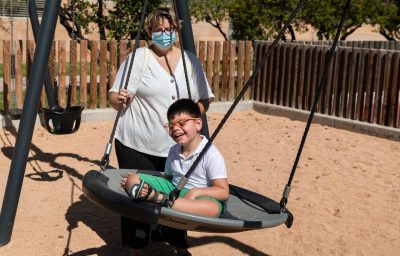Child with disability in swing with mother pushing, wears blue mask of pandemic coronavirus protection.