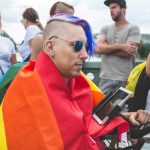 Hipster man in a rainbow flag in a wheelchair with a tablet in a crowd