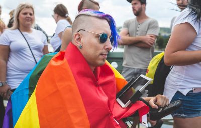 Hipster man in a rainbow flag in a wheelchair with a tablet in a crowd