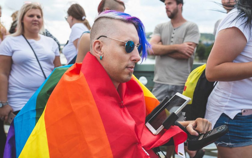 Hipster man in a rainbow flag in a wheelchair with a tablet in a crowd