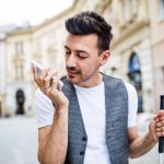 Young blind man with smartphone walking on street in city, using voice assistant