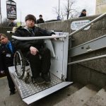 Worker of the underground is helping to the disabled person in a wheelchair to move around on the stairs with a special lifting device.