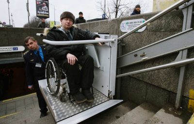 Worker of the underground is helping to the disabled person in a wheelchair to move around on the stairs with a special lifting device.