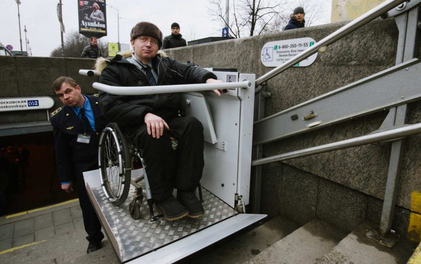 Worker of the underground is helping to the disabled person in a wheelchair to move around on the stairs with a special lifting device.