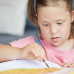 Child with down syndrome painting with paintbrush at the table at home