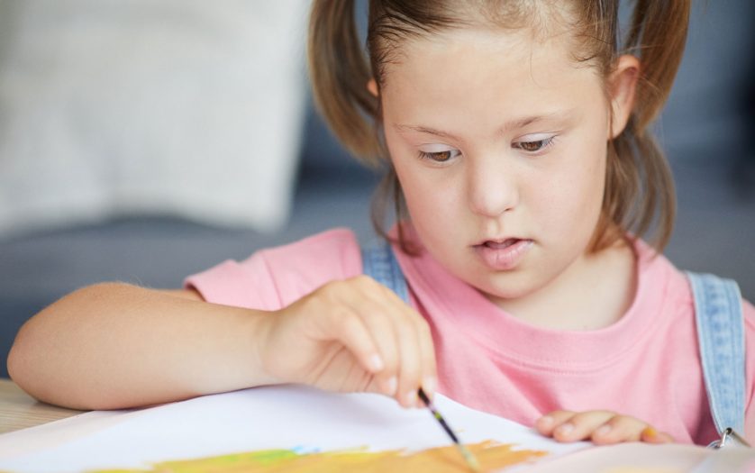 Child with down syndrome painting with paintbrush at the table at home