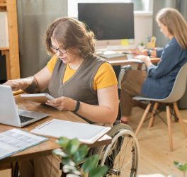 Young woman in wheelchair in front of laptop and networking while her colleague working behind
