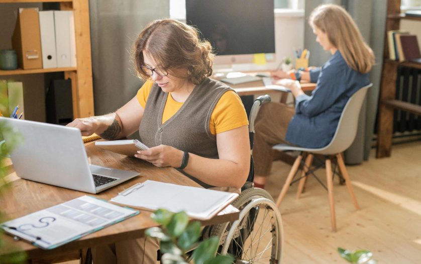 Young woman in wheelchair in front of laptop and networking while her colleague working behind