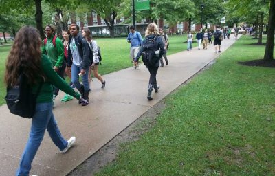 A campus sidewalk is filled with male and female students walking to and from class