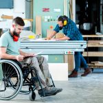 Worker in wheelchair in a carpenter`s workshop with his colleague