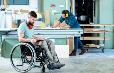 Worker in wheelchair in a carpenter`s workshop with his colleague