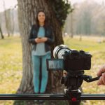 Video shooting at the park: video camera and operator hand on foreground and female model standing in the background