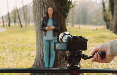 Video shooting at the park: video camera and operator hand on foreground and female model standing in the background
