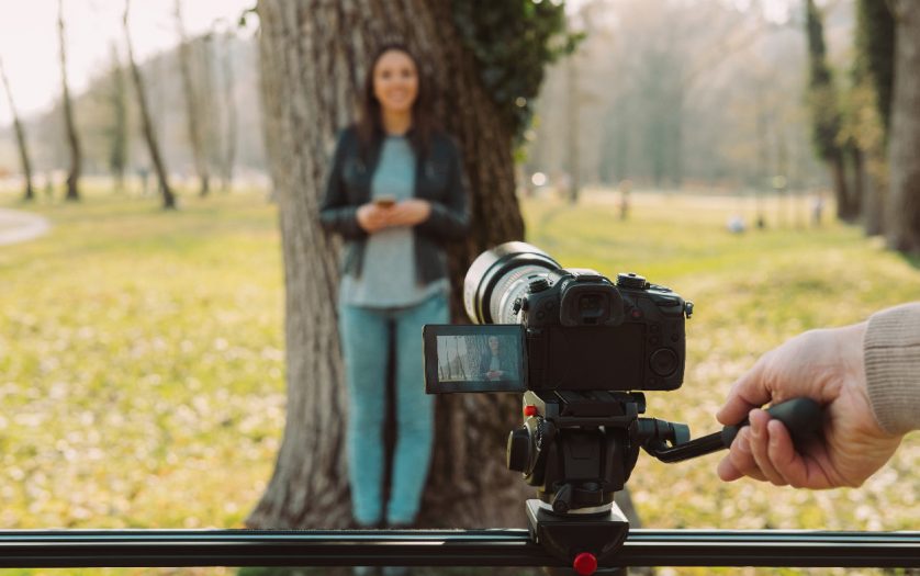 Video shooting at the park: video camera and operator hand on foreground and female model standing in the background