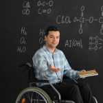 Male teacher with books in wheelchair near chalkboard