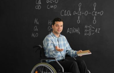 Male teacher with books in wheelchair near chalkboard