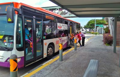 A bus driver lowers the ramp at the rear door for the helper to push a wheelchair bound passenger up onto the bus