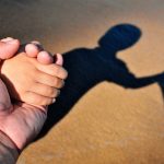 Closeup of father and kid holding hands with their shadows in brown sand background.