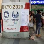 A wrapped column in front of Chiba Station, Japan with Tokyo Olympics & Paralympics logos & a poster with advice on coronaviurs safety measures. People wear face masks.