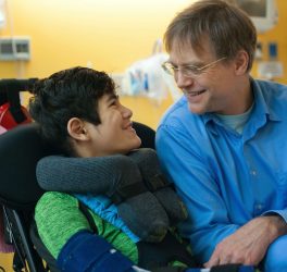 Smiling father sitting next to disabled son in wheelchair by hospital bed, talking together