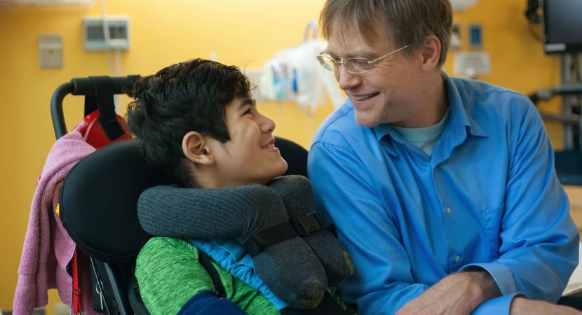 Smiling father sitting next to disabled son in wheelchair by hospital bed, talking together