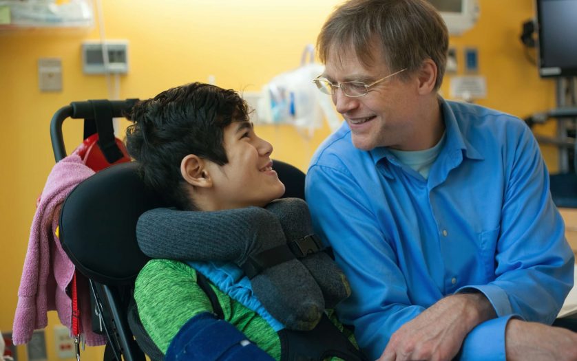 Smiling father sitting next to disabled son in wheelchair by hospital bed, talking together