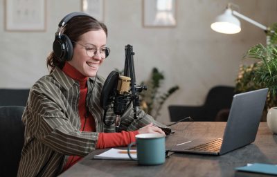 Young reporter in headphones sitting at the table looking at monitor of laptop and talking online in the studio