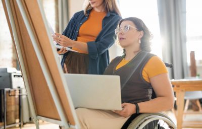 Young woman in casualwear helping woman in wheelchair with presentation while standing in front of whiteboard in office