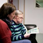 Caregiver looking at man with cognitive disability reading magazine at home