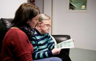 Caregiver looking at man with cognitive disability reading magazine at home