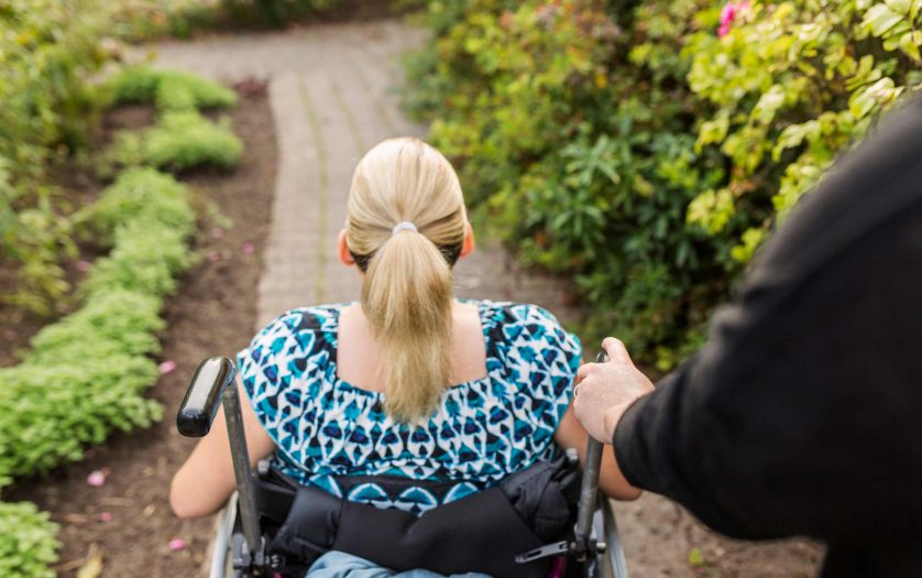 Disabled woman in wheelchair with assistant walking in garden