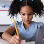 schoolgirl using digital tablet while writing on book at desk in classroom