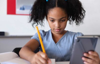 schoolgirl using digital tablet while writing on book at desk in classroom