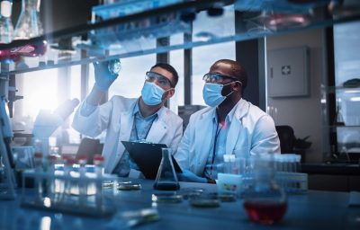 Two Scientists Wearing Face Masks use Microscope, Analyse Sample in Petri Dish, Discuss Innovative Technology
