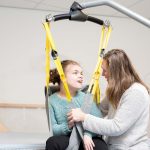 A disabled girl being lifted into a wheelchair with help from a special lift operated by a care assistant.