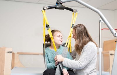 A disabled girl being lifted into a wheelchair with help from a special lift operated by a care assistant.