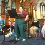 Four people sit on a church platform with one person standing at a lecturn with a microphone. The person standing has short green hair and wears green overalls.