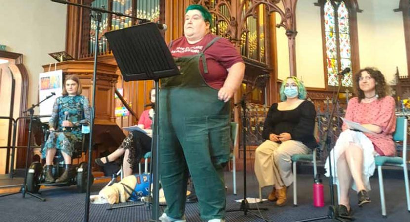 Four people sit on a church platform with one person standing at a lecturn with a microphone. The person standing has short green hair and wears green overalls.