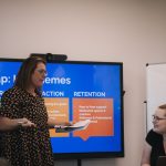 a photo of network members Melissa Hale (left) and Jessi Hooper (right) facilitating a Building Inclusive Sport Clubs workshop in a meeting room. Melissa is standing and speaking holding notes and a pen. Jessi is sitting, listening and smiling. Behind the two is a television screen with a presentation.