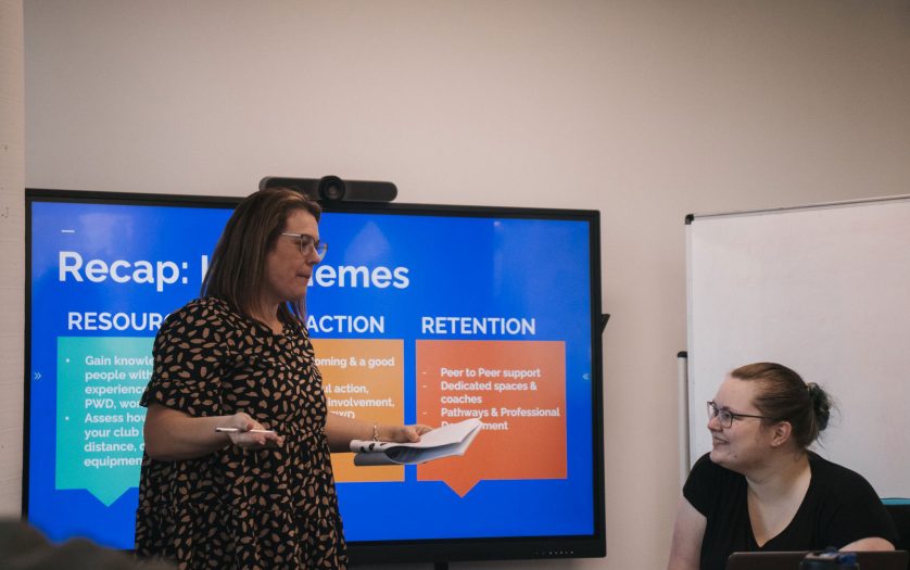 a photo of network members Melissa Hale (left) and Jessi Hooper (right) facilitating a Building Inclusive Sport Clubs workshop in a meeting room. Melissa is standing and speaking holding notes and a pen. Jessi is sitting, listening and smiling. Behind the two is a television screen with a presentation.