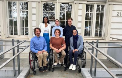Six athletes, three standing in the back and three in wheelchairs at the front, pose for a photo in front of the IPC headquarters.