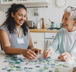 senior lady having fun plying puzzle game at kitchen table with black female worker