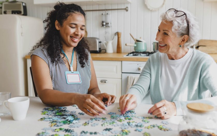 senior lady having fun plying puzzle game at kitchen table with black female worker